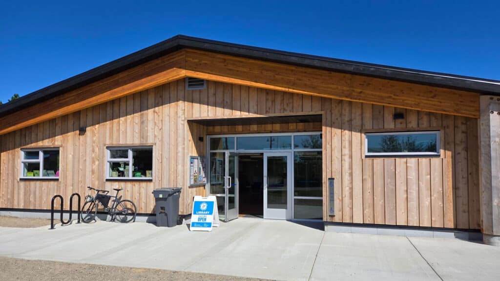 The front facade of the new Masset library. It is a wood building with a set of double glass doors that are open. There is a sandwich board that says OPEN with a black metal bike rack.